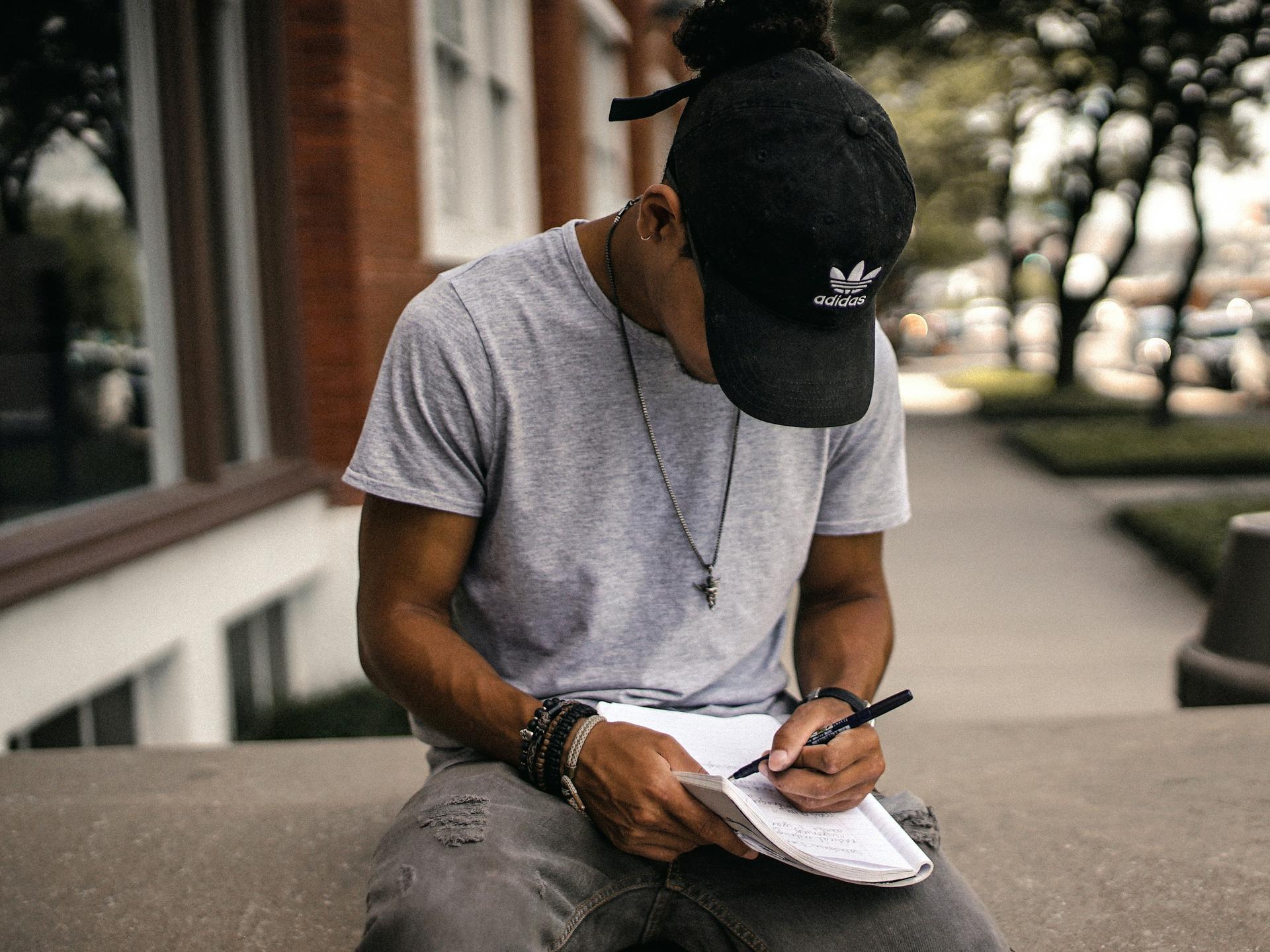 Man writing in notebook, sitting on concrete divider in daytime, looking down, unemployed