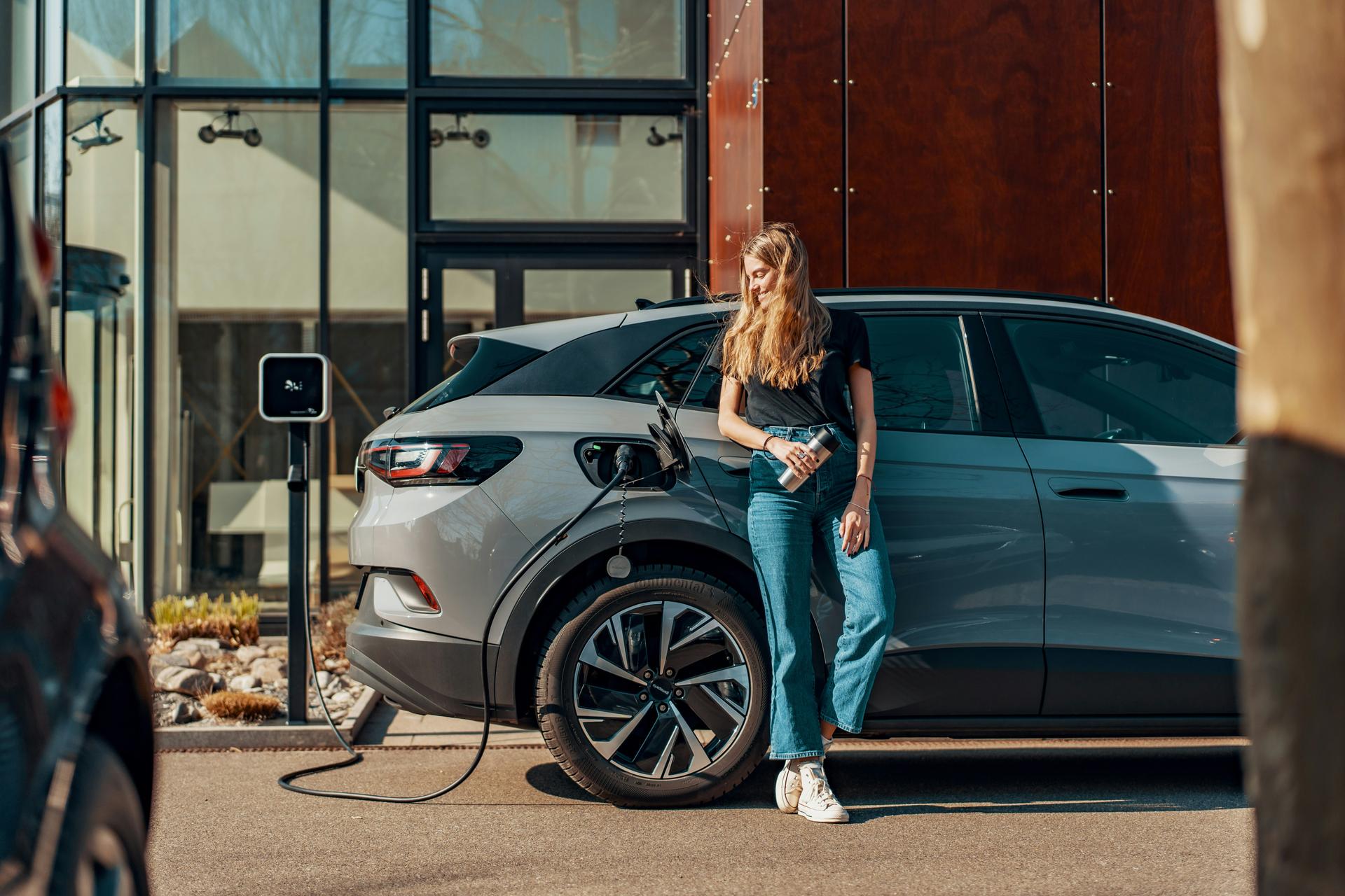 a young woman leans on here electric vehicle while it charges
