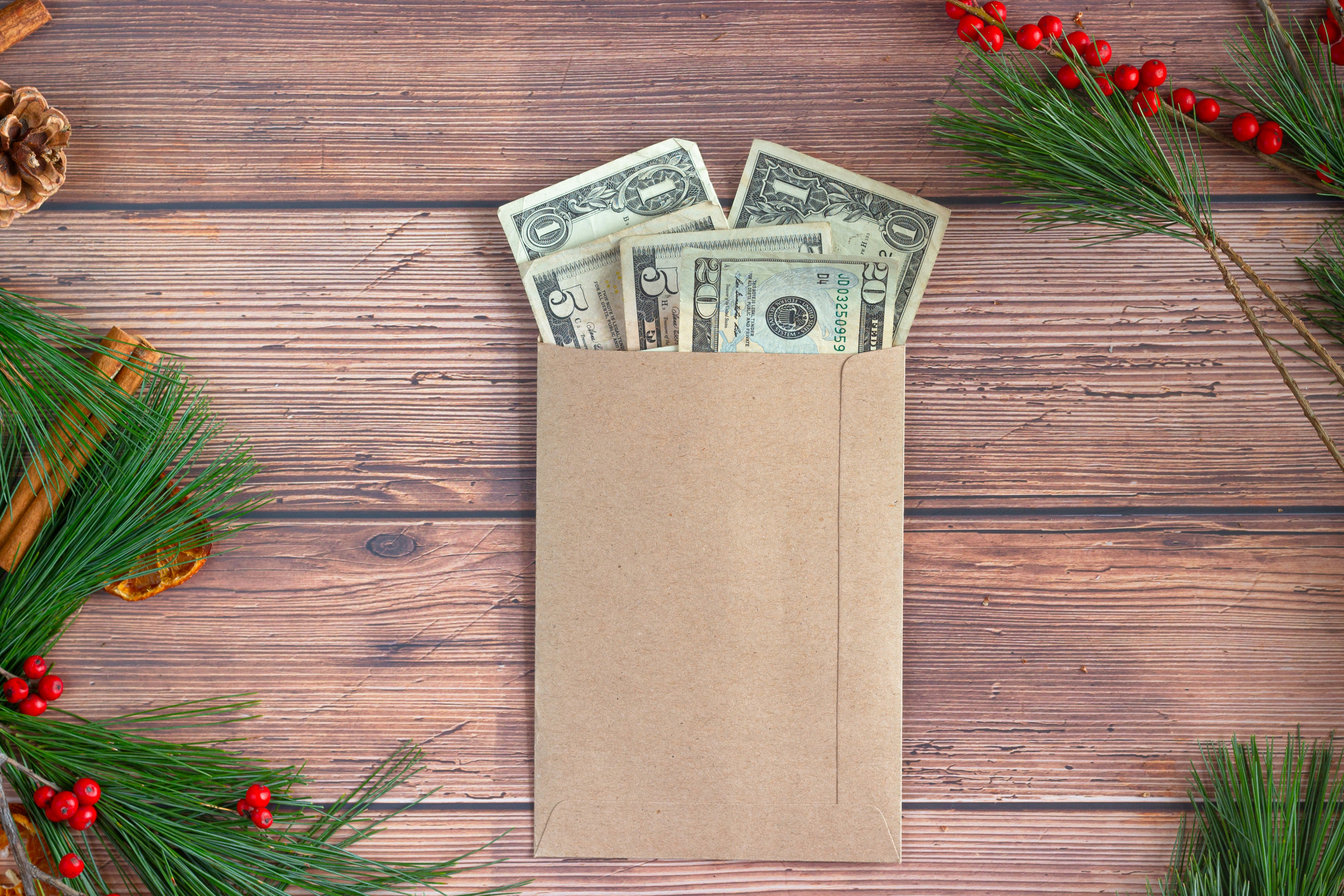 A brown envelope of American money on a wood surface surrounded by festive spruce needles, cinnamon, and red berries.