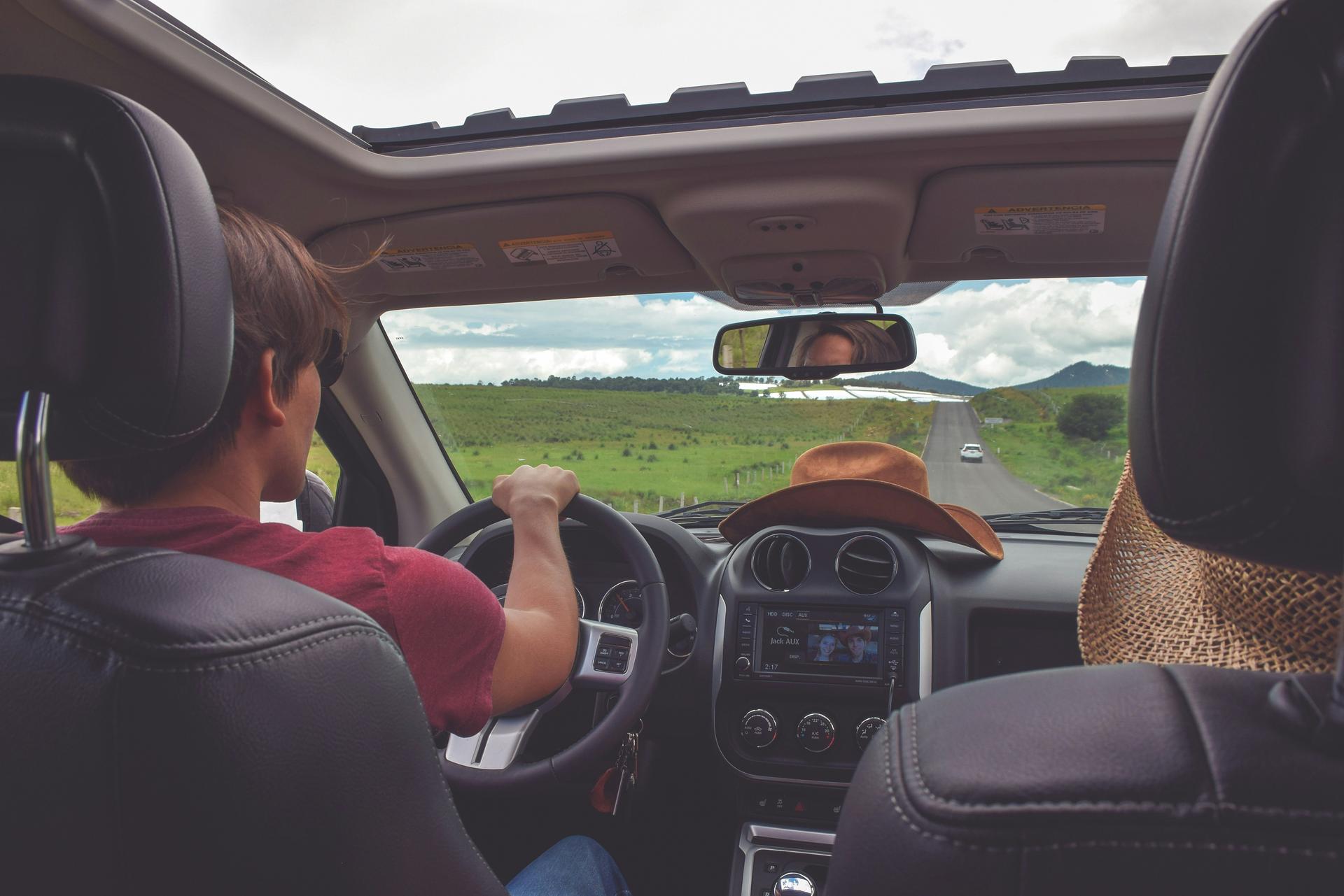 man driving car on rural road with cowboy hat on dash, woman in passenger seat with woven sun hat