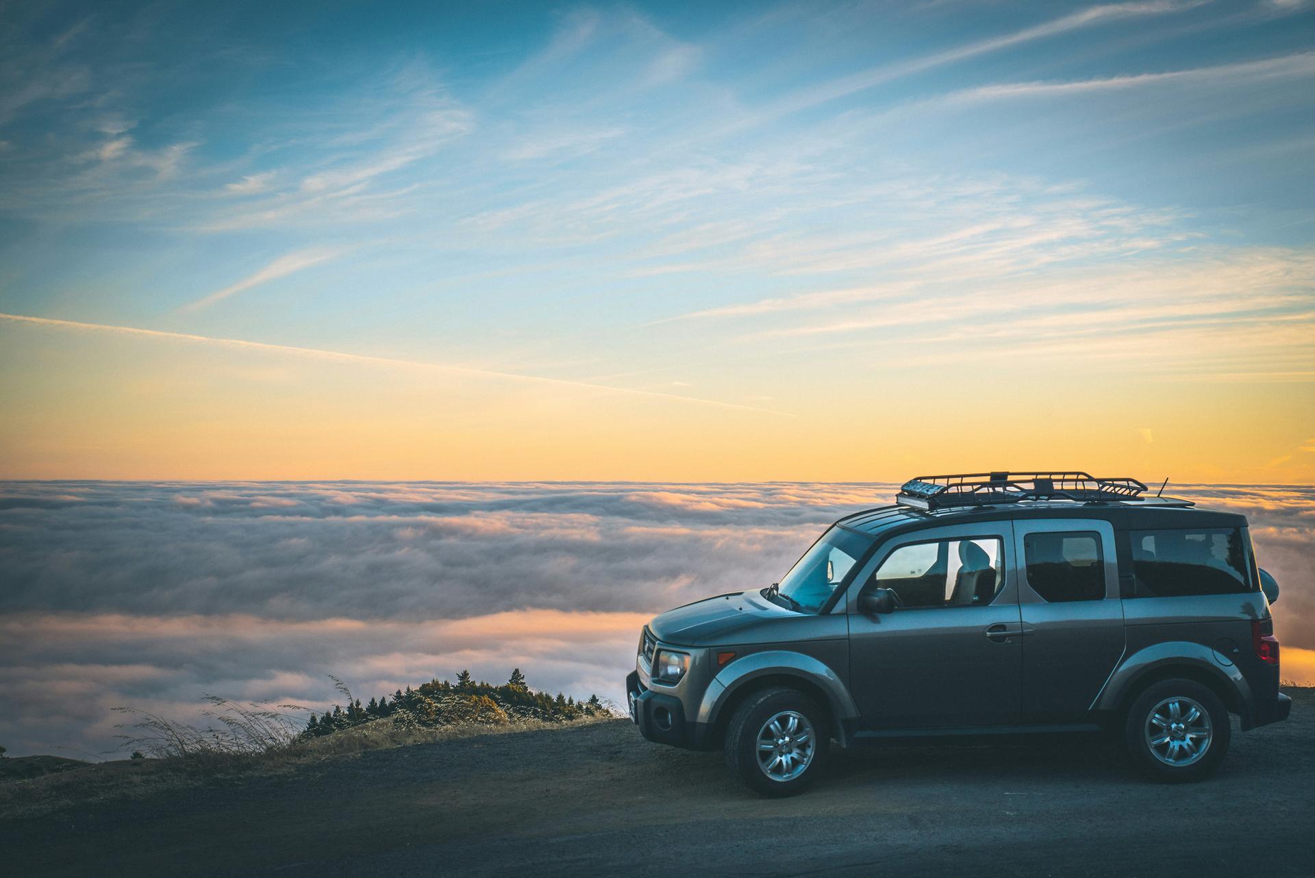 silver van-SUV high up on a grassy cliff overlooking forest and clouds
